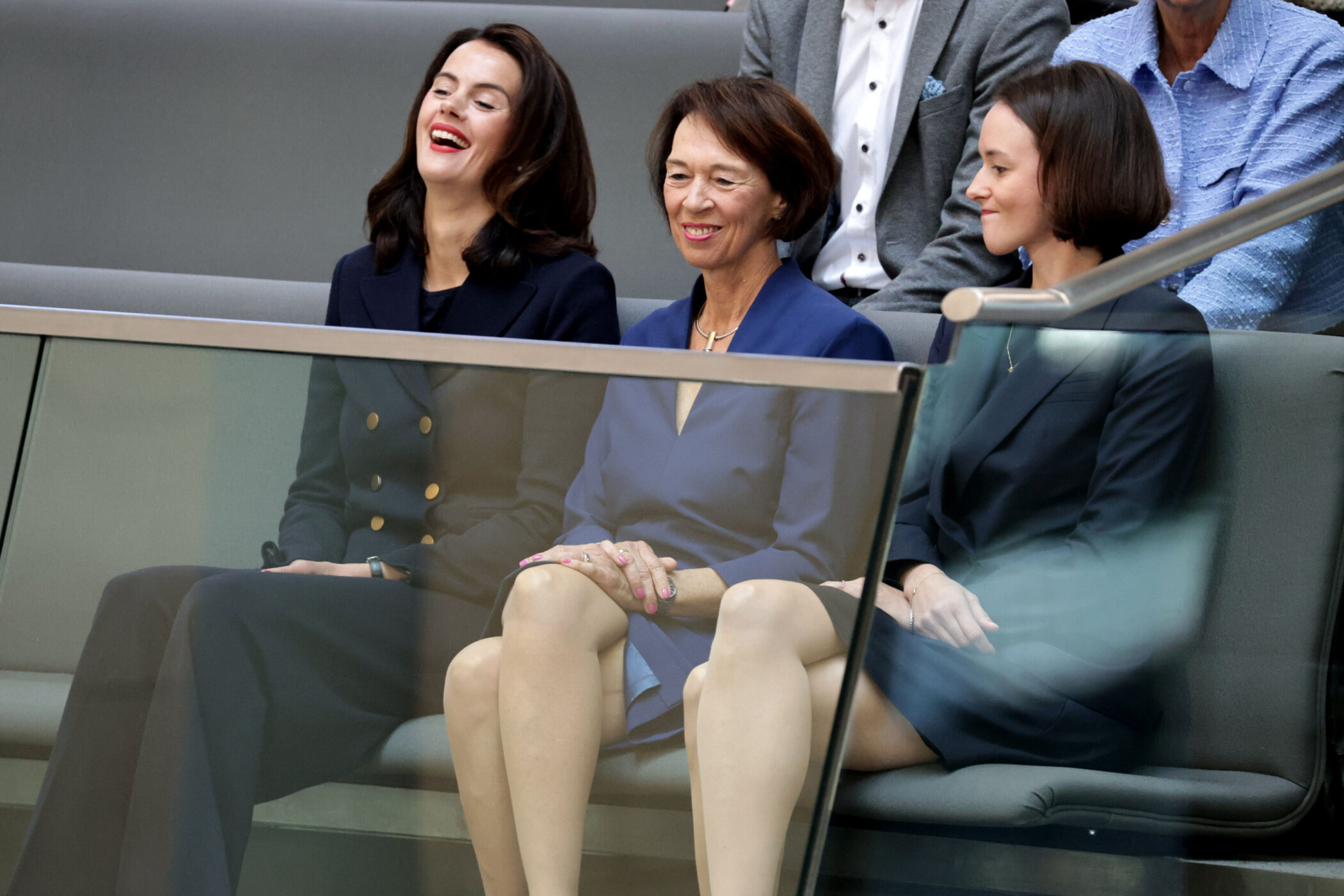 Drei Personen in Anzugjacken sitzen auf einer Bank, eine Person lacht.