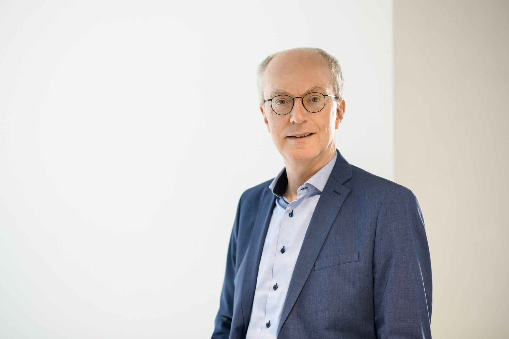 Mann mit Brille im blauen Anzug und hellblauem Hemd vor weißem Hintergrund.