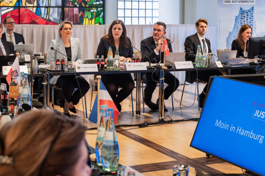 Sieben Personen sitzen an einem Konferenztisch mit Laptops und Getränken. Bildschirm zeigt „Moin in Hamburg“.