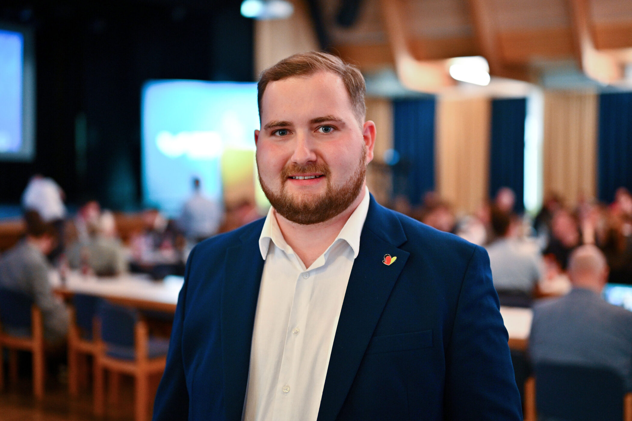 Ein Mann in Anzug steht in einem Raum mit Leuten im Hintergrund.