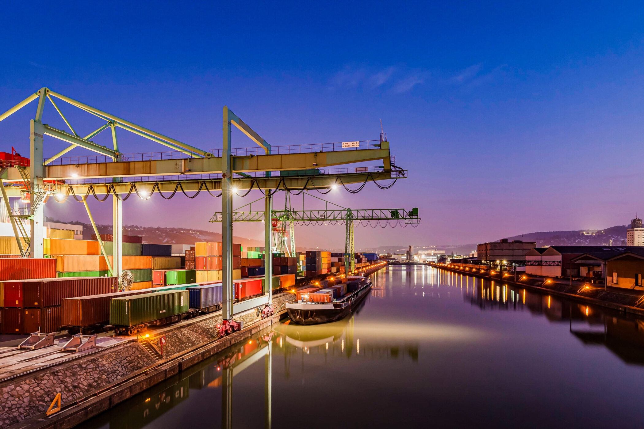 Containerkräne und Frachtschiff an einem Wasserkanal bei Dämmerung.