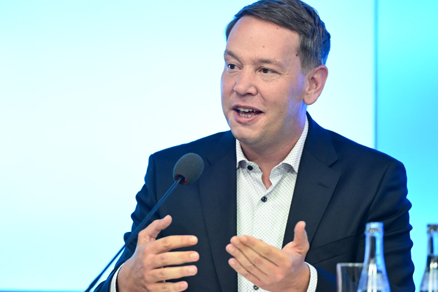 Mann im Anzug spricht gestikulierend ins Mikrofon, vor hellem Hintergrund.
