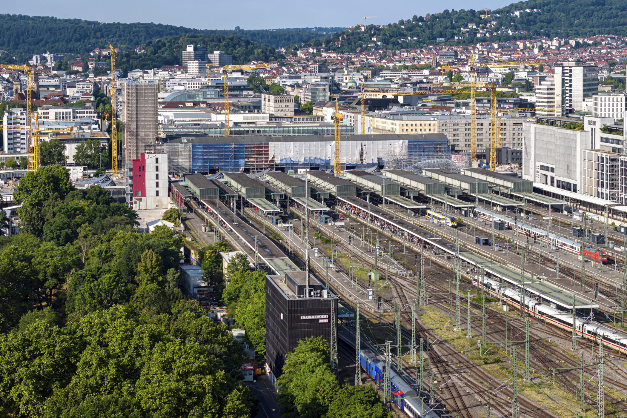 Hauptbahnhof mit vielen Gleisen, Zügen und Baukränen, im Hintergrund Stadt und Hügel.