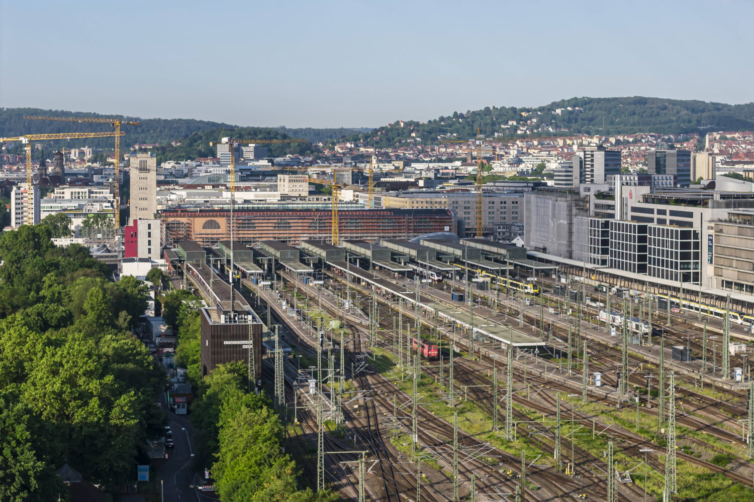 Bahnhof mit vielen Gleisen, Gebäuden im Hintergrund und bewaldetem Bereich links.