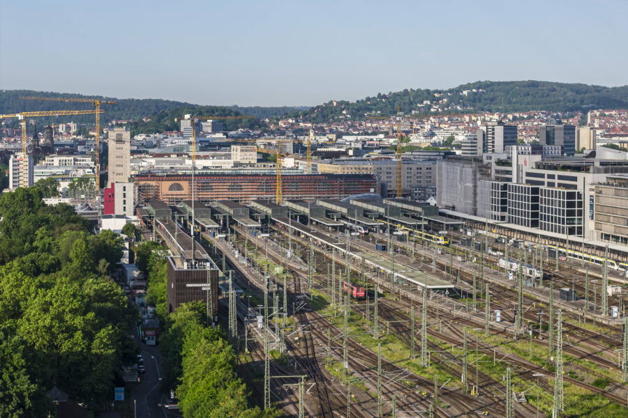 Bahnhof mit vielen Gleisen, Gebäuden im Hintergrund und bewaldetem Bereich links.