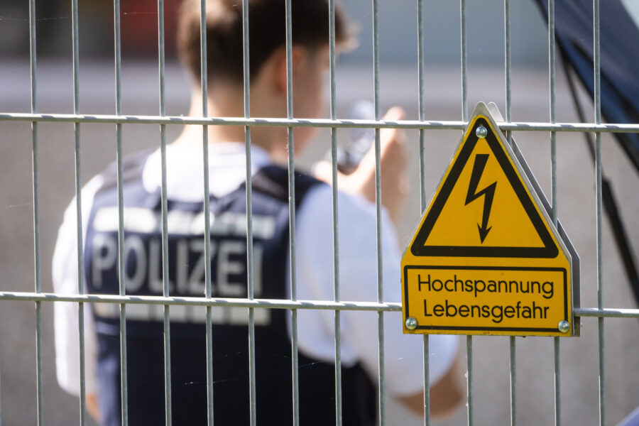 Polizist hinter Zaun mit Schild „Hochspannung Lebensgefahr“.