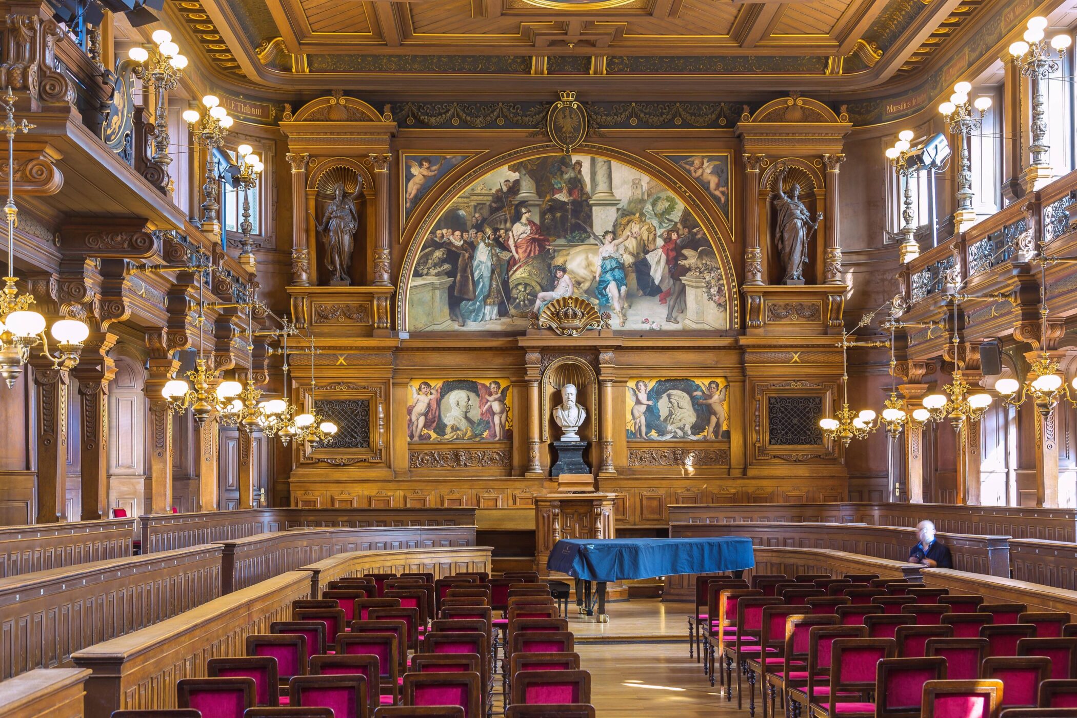 Prunkvoller Saal mit Holzvertäfelung, Gemälden, Kronleuchtern und roten Stühlen.