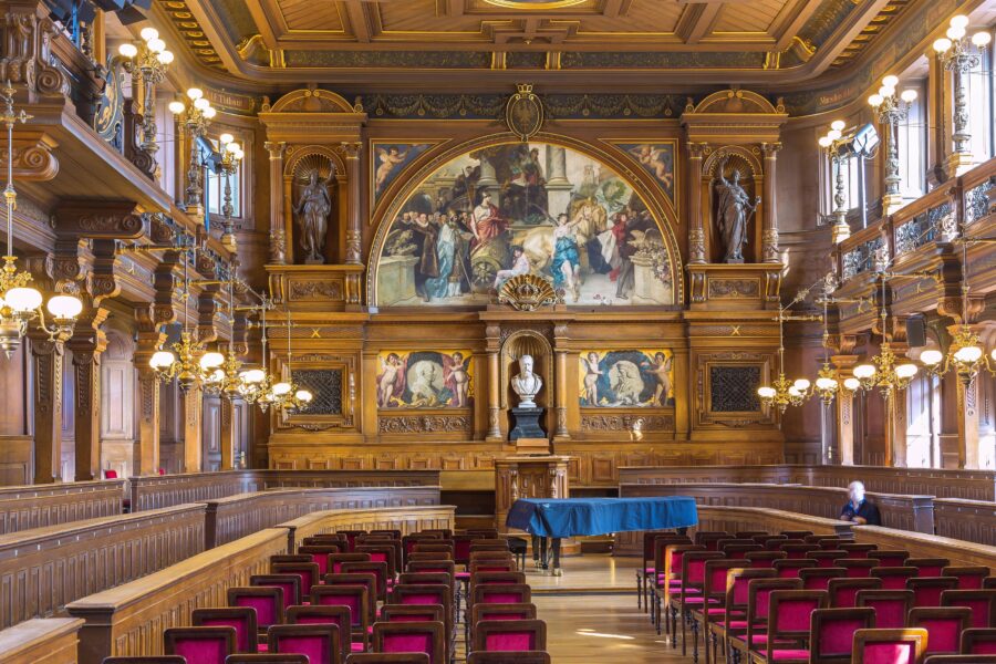 Prunkvoller Saal mit Holzvertäfelung, Gemälden, Kronleuchtern und roten Stühlen.