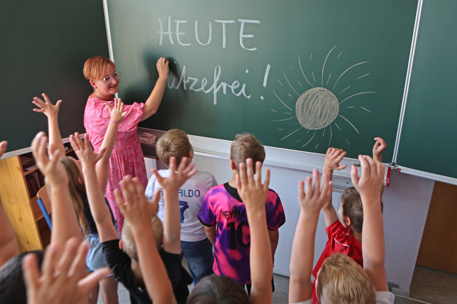 Person schreibt "HEUTE hitzefrei!" und Sonne an Tafel, Kinder heben Hände.