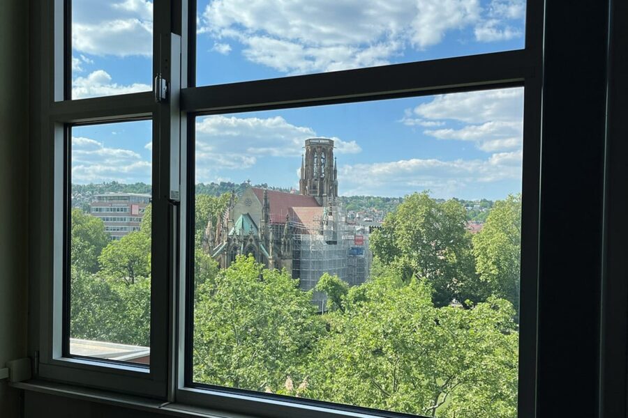 Blick aus einem Fenster auf eine Kirche und Bäume bei blauem Himmel mit Wolken.