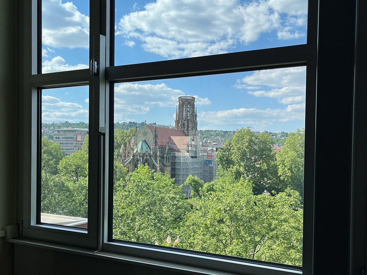 Blick aus einem Fenster auf eine Kirche und Bäume bei blauem Himmel mit Wolken.