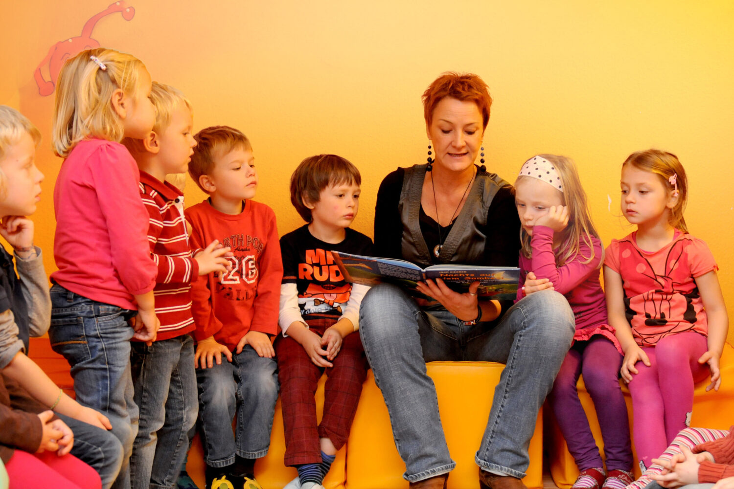 Eine Person liest mehreren Kindern ein Buch vor. Hintergrund gelb.