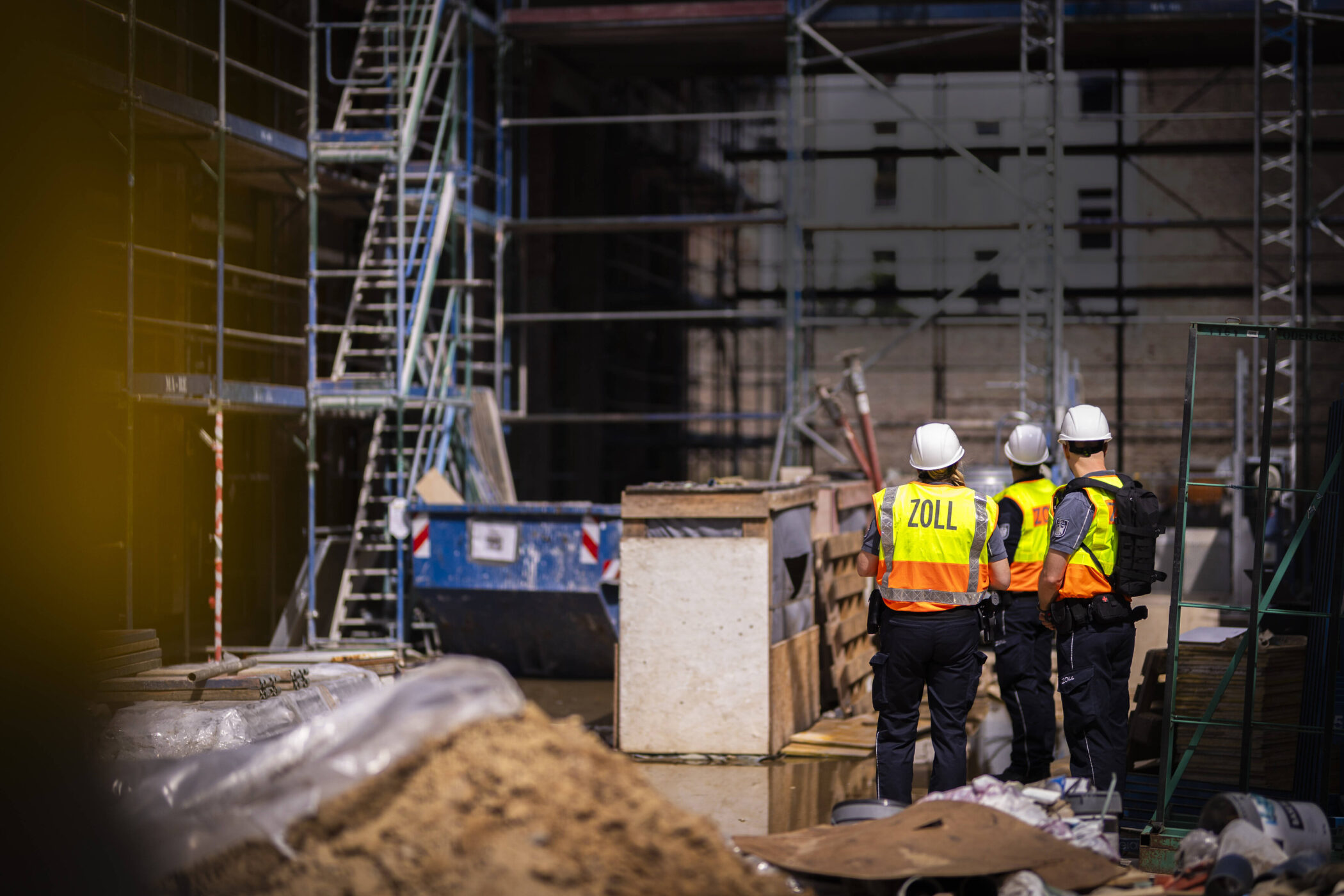 Drei Personen mit Helmen und Warnwesten auf Baustelle, Rückansicht.