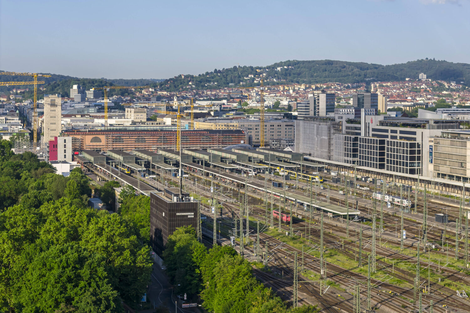 Weitläufige Bahnanlage mit Zügen, Gebäuden und Kränen, im Hintergrund Hügel.