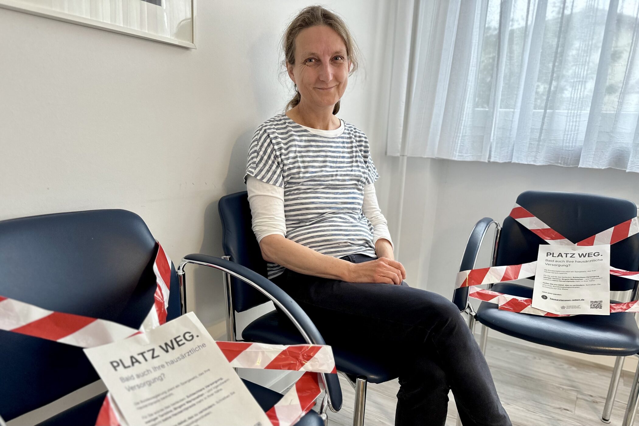 Frau sitzt in einem Wartezimmer, zwei Stühle mit Absperrband markiert.