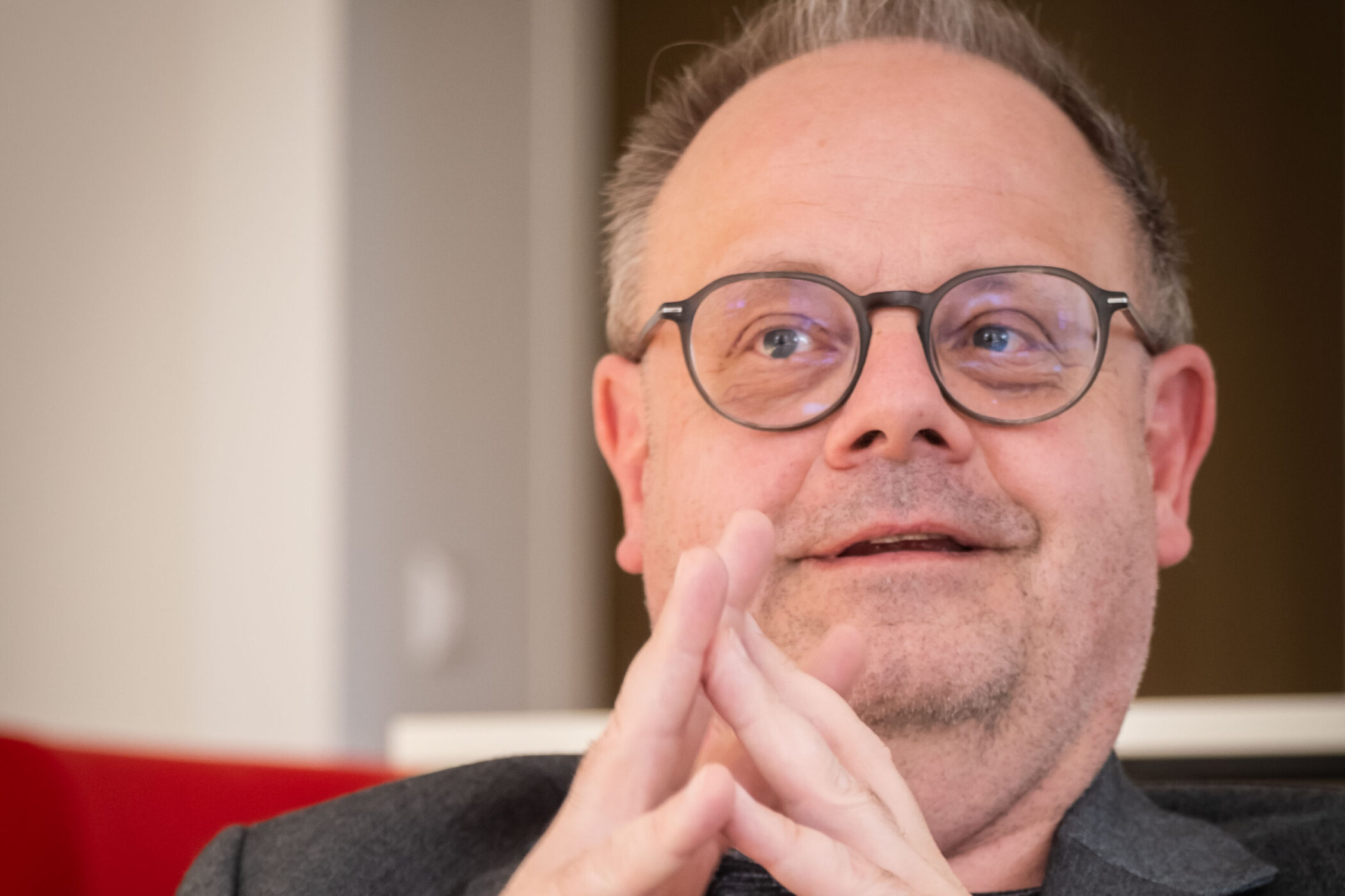 Mann mit Brille und gefalteten Händen, im Hintergrund rote Couch.
