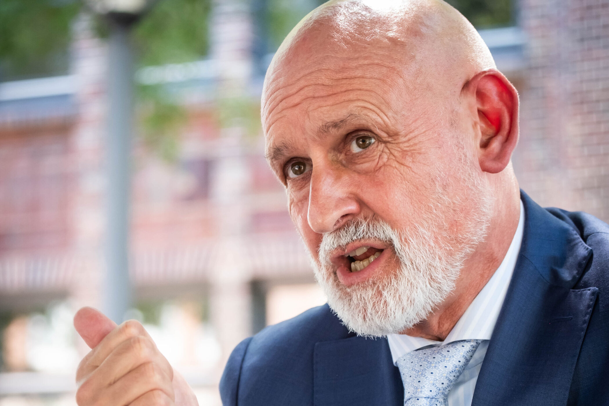 Älterer Mann in Anzug spricht gestikulierend, unscharfer Hintergrund.