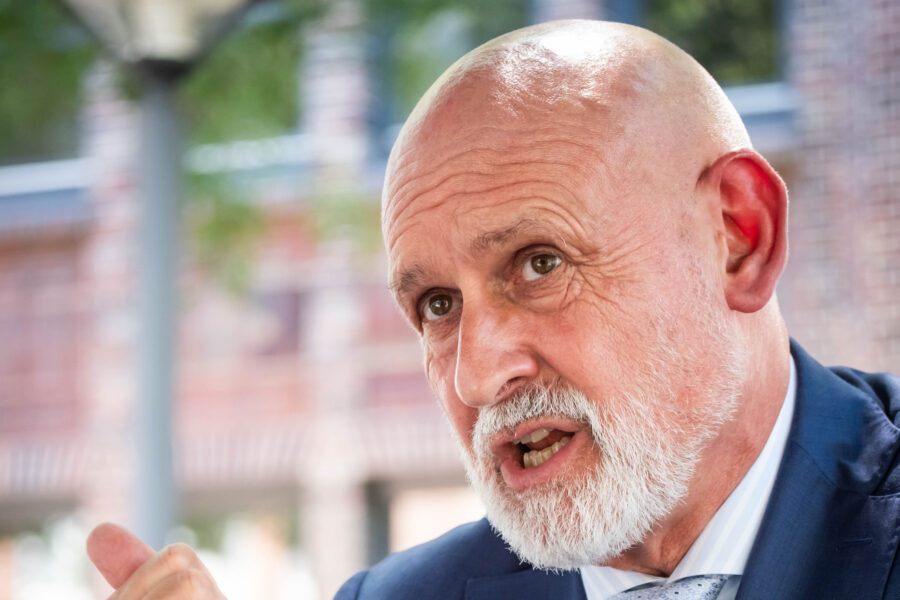 Älterer Mann mit Bart spricht, Hintergrund aus Ziegelwänden und Grün.