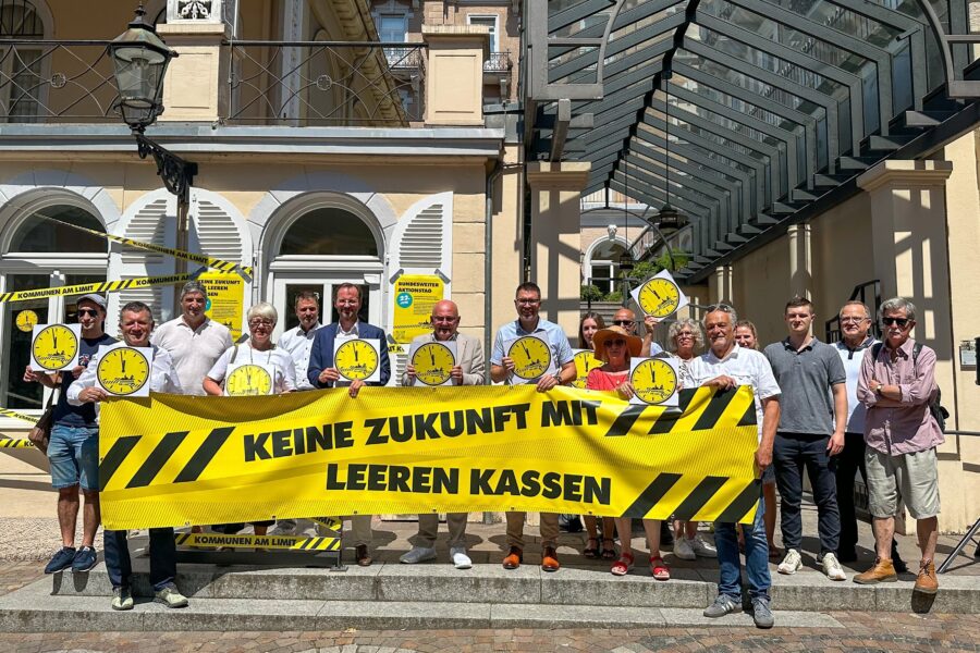 15 Personen stehen mit gelbem Banner "KEINE ZUKUNFT MIT LEEREN KASSEN" vor Gebäude.
