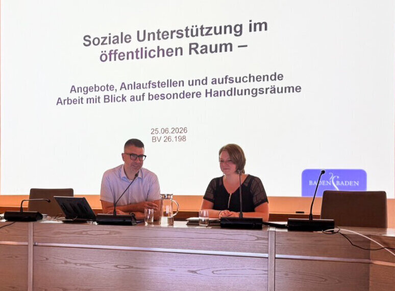 Zwei Personen sitzen an einem Tisch vor einer Präsentation mit dem Thema „Soziale Unterstützung“.