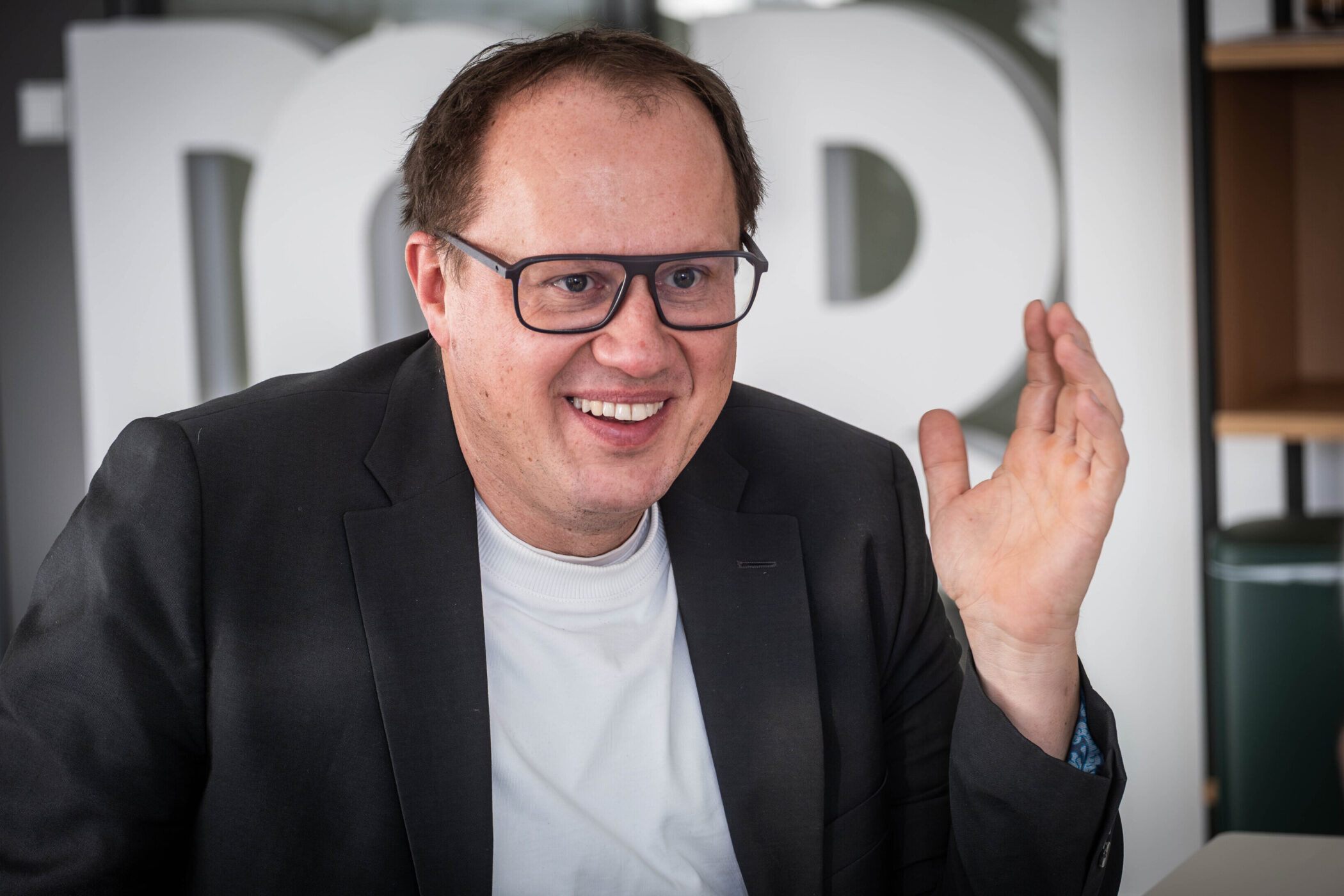 Person mit Brille, in schwarzem Sakko, hebt eine Hand beim Sprechen.