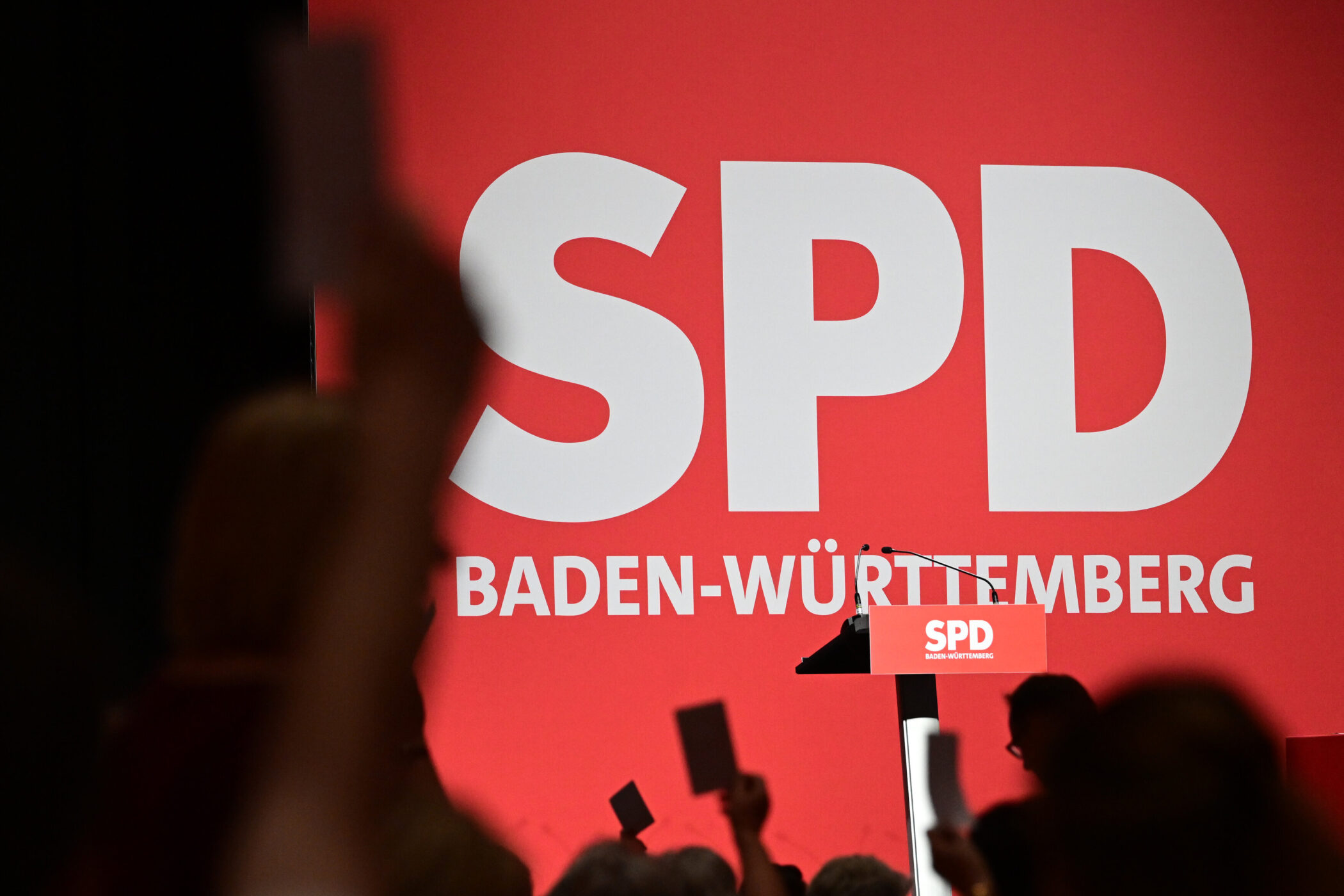 Roter Hintergrund mit „SPD Baden-Württemberg“ und Pult mit SPD-Logo, Hände halten Karten hoch.