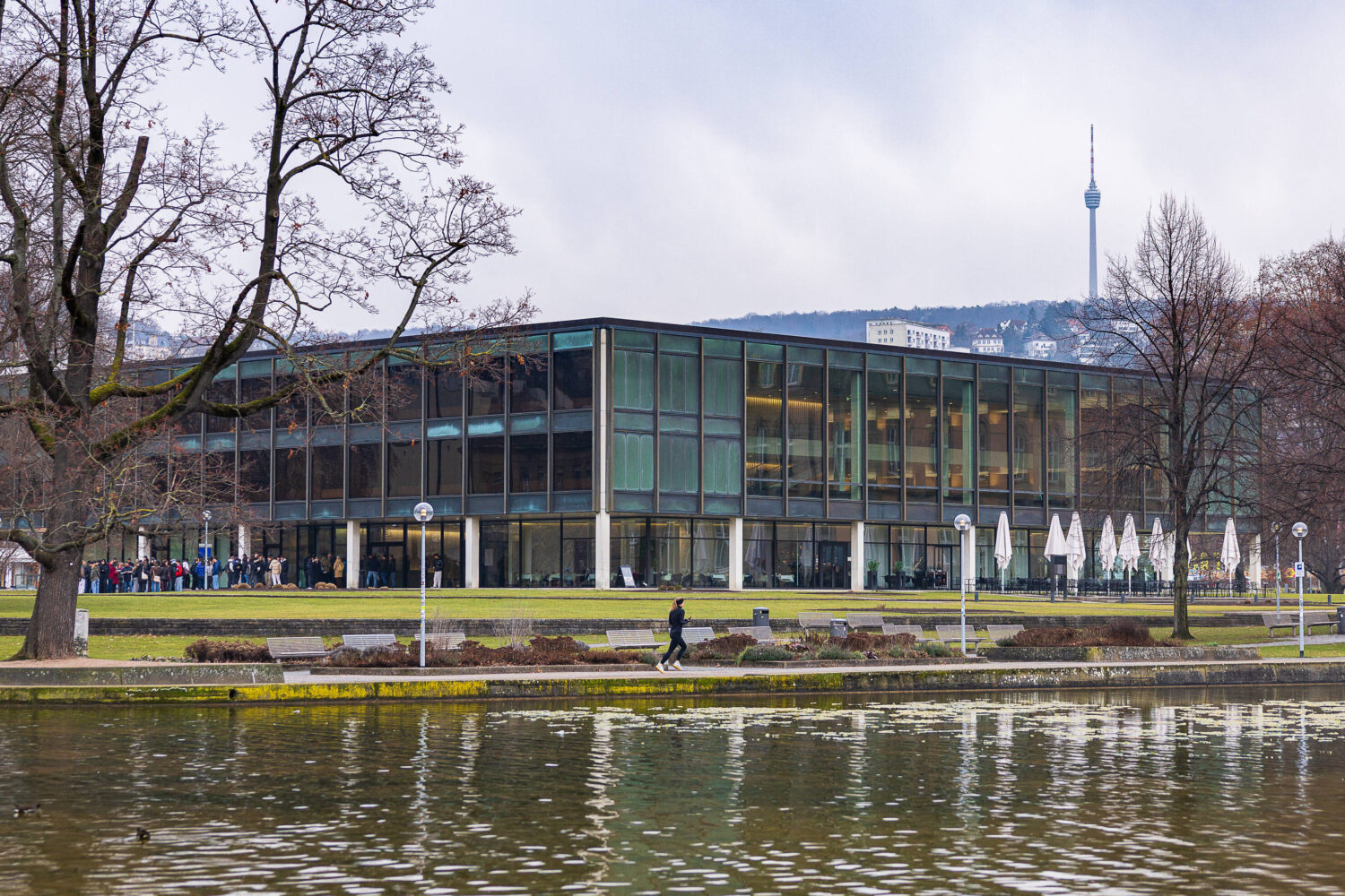 Modernes Gebäude mit Glasfassade und Bäumen am Ufer eines Gewässers, Personen gehen entlang.
