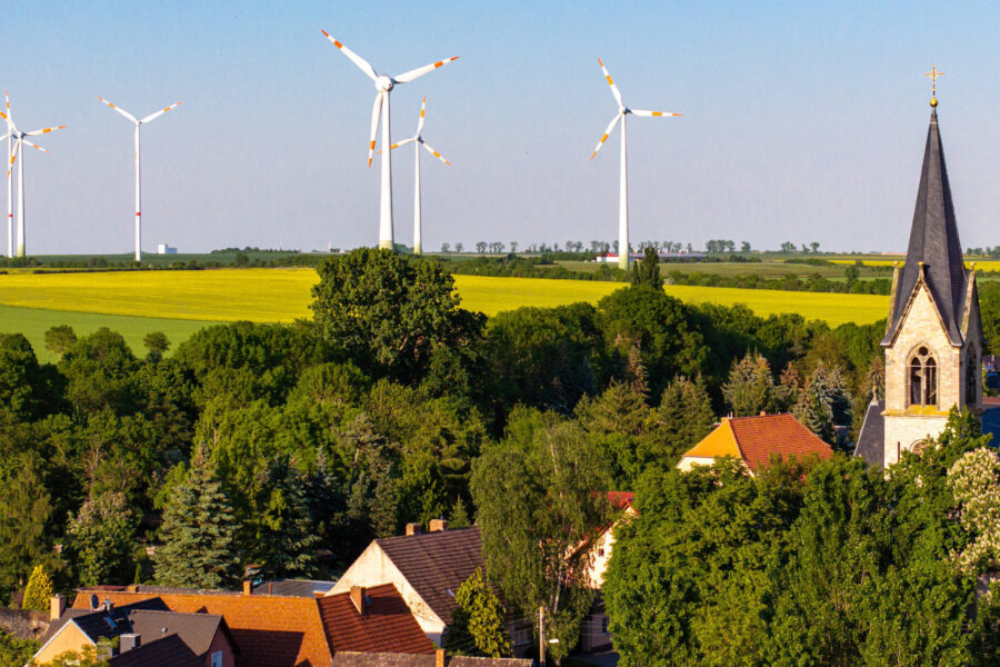 Kirchturm, Häuser und Bäume im Vordergrund, Windräder im Hintergrund.
