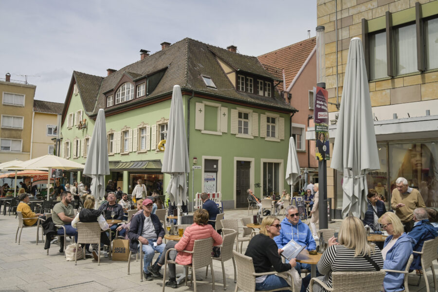 Menschen sitzen bei Sonnenschein an Tischen vor einem grünen Haus in der Stadt.