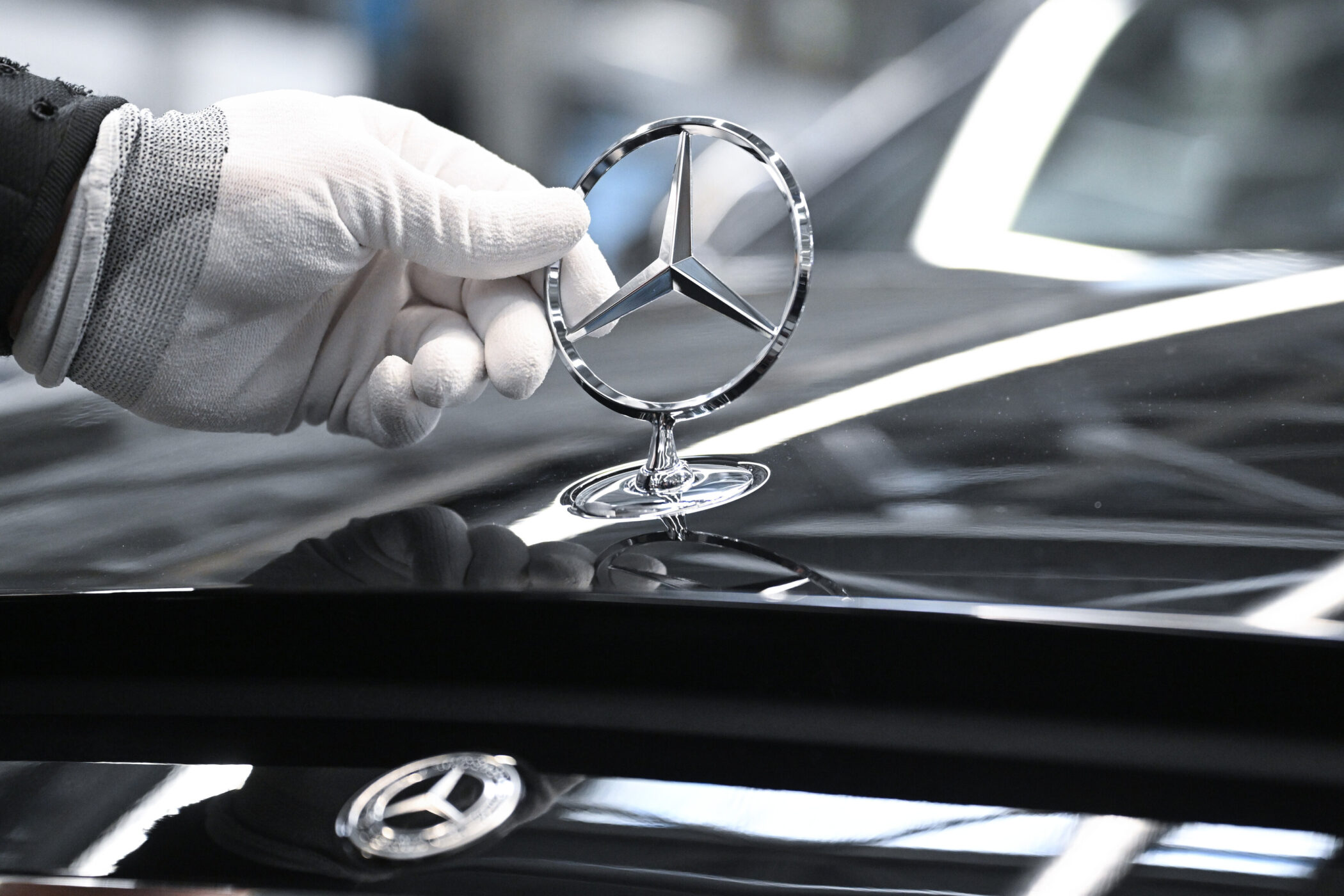 Hand mit Handschuh hält ein Mercedes-Emblem auf einer glänzenden Motorhaube.