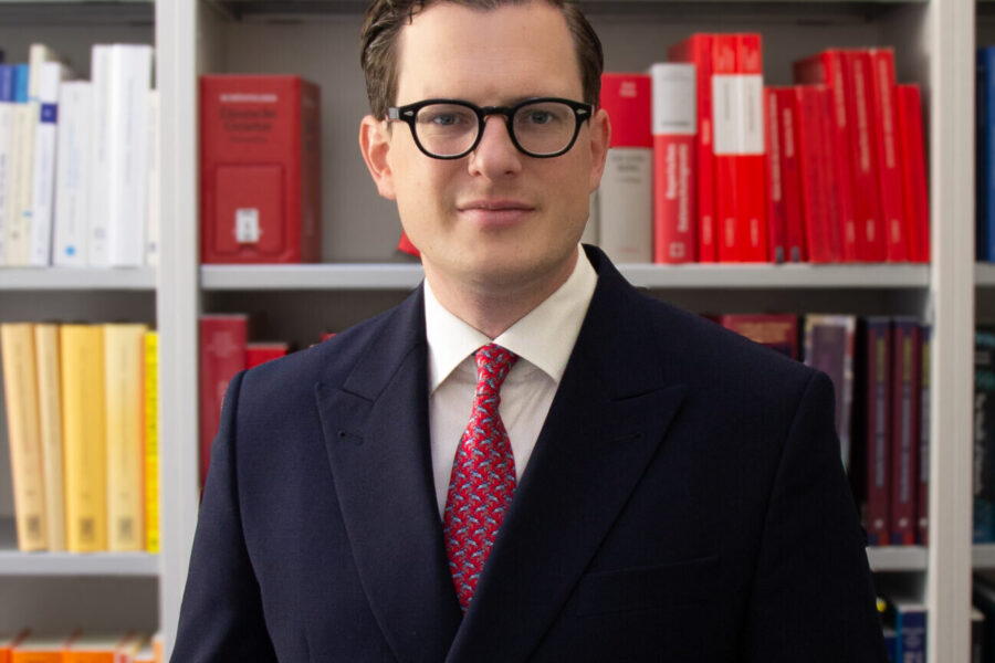 Mann in Anzug vor Bücherregal mit roten und weißen Büchern.