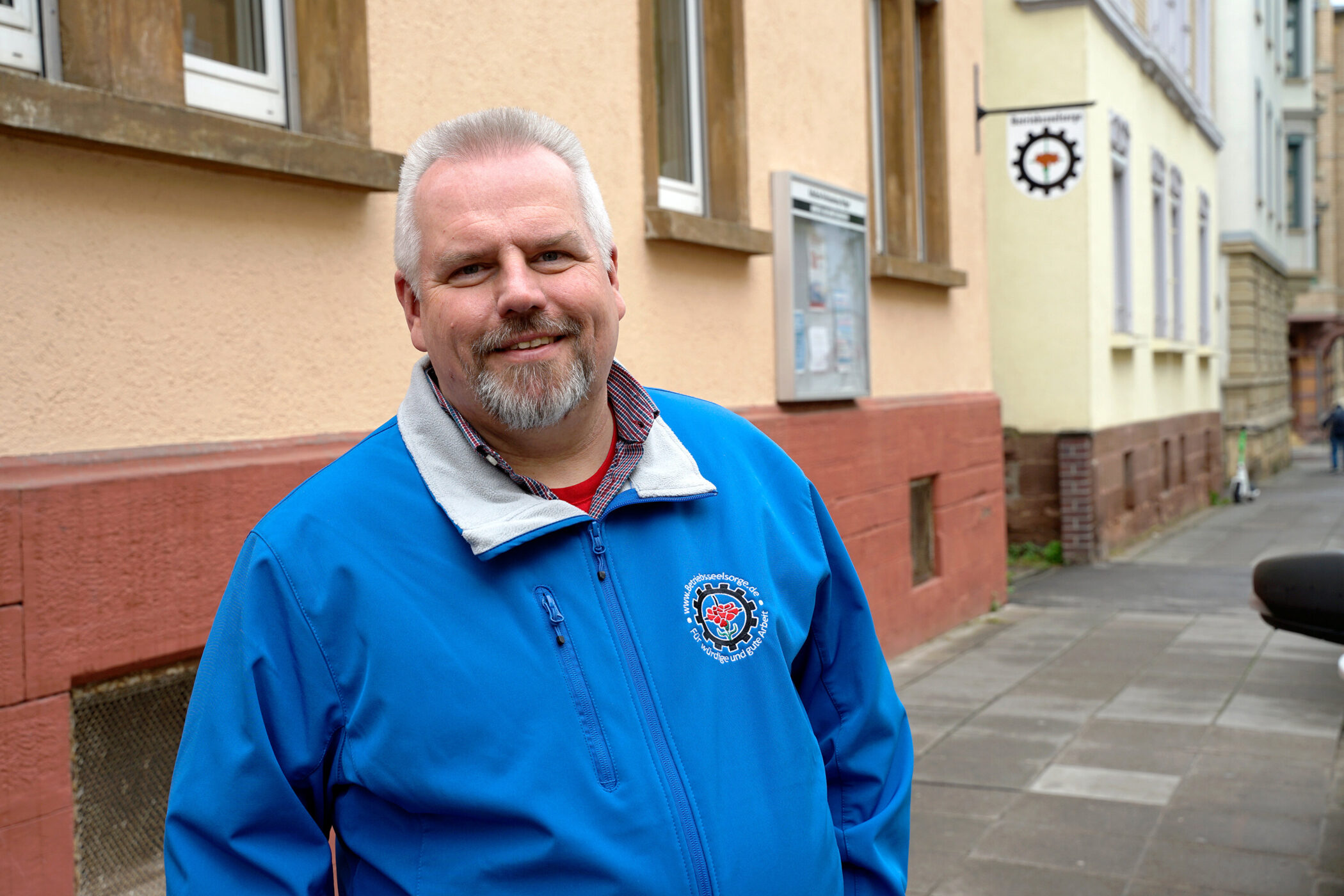 Mann in blauer Jacke lächelt, steht vor einem Gebäude auf dem Bürgersteig.