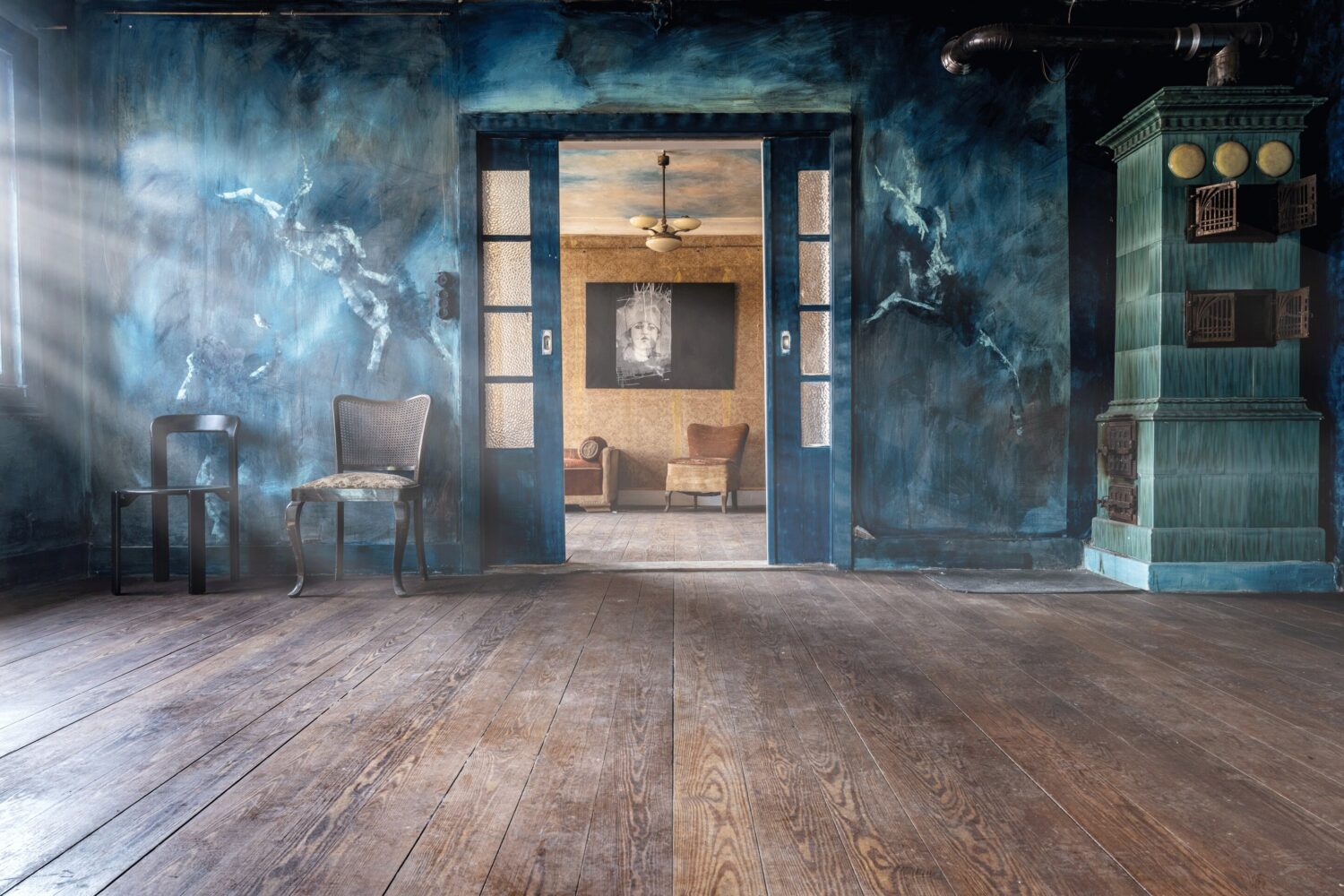 Holzfußboden, blaue Wand, zwei Stühle, Kamin, geöffnete Schiebetür zeigt Raum im Hintergrund.