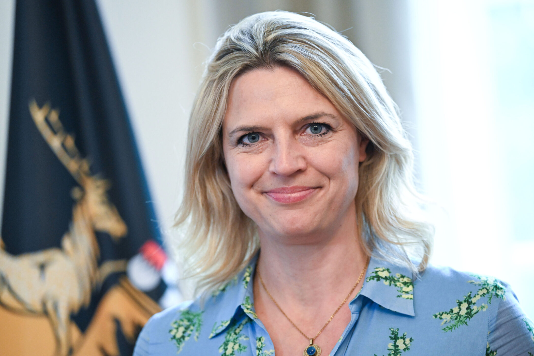 Lächelnde Frau mit blonden Haaren, blauer Bluse, Fahne im Hintergrund.