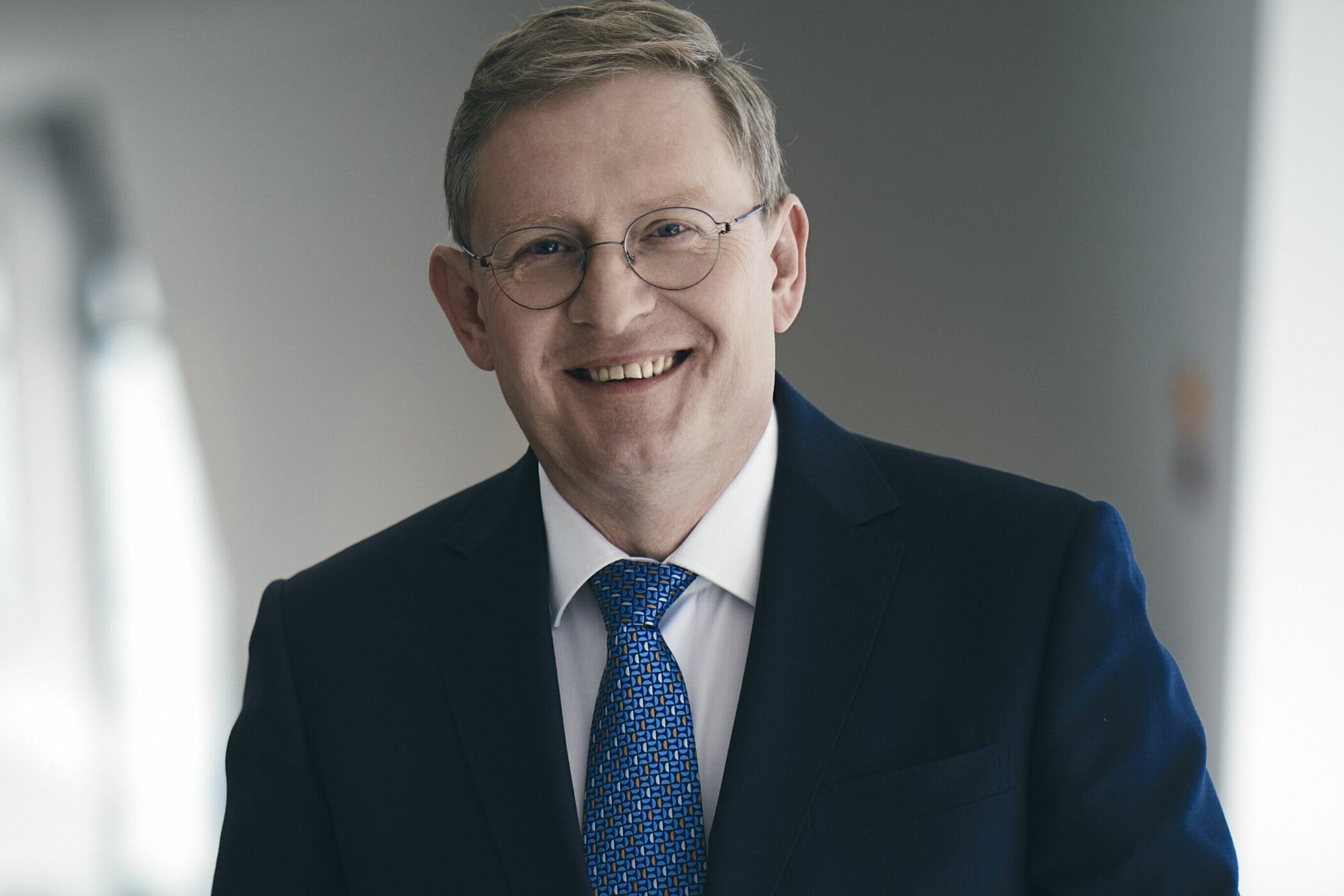 Mann in Anzug und Krawatte, lächelnd, mit Brille, vor grauem Hintergrund.
