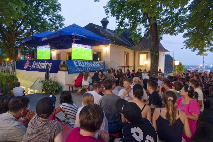 Menschen sitzen draußen und schauen Fußball auf Bildschirmen unter Sonnenschirmen.