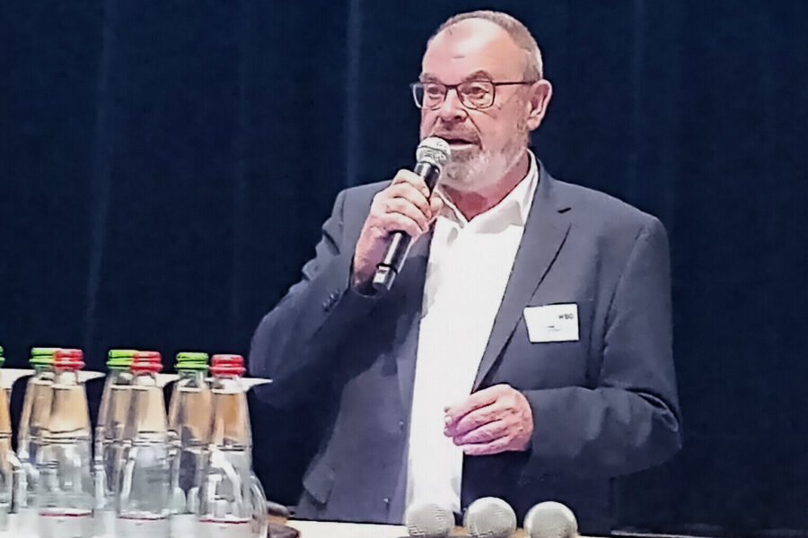 Mann in Anzug spricht ins Mikrofon, Wasserflaschen im Vordergrund.