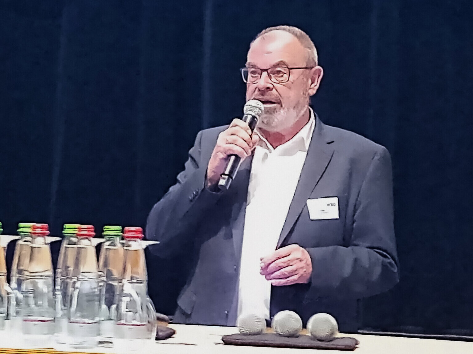 Mann in Anzug spricht ins Mikrofon, Wasserflaschen im Vordergrund.