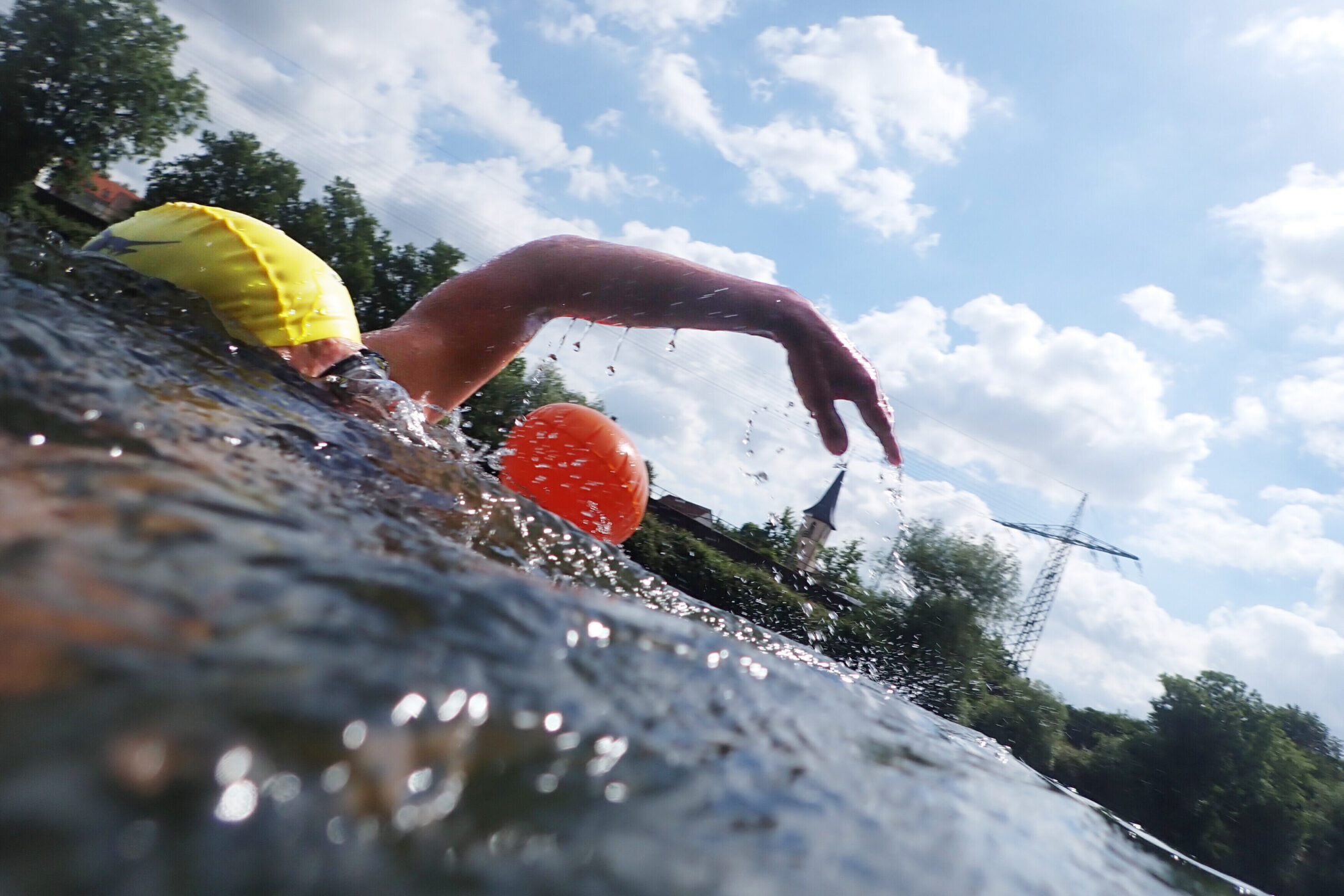 Person schwimmt im Wasser mit gelber Badekappe, blauer Himmel und Wolken im Hintergrund.