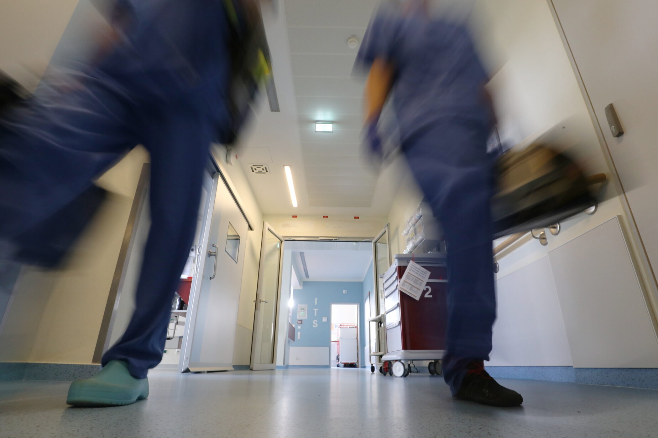 Zwei Personen in Krankenhauskleidung gehen fluraufwärts. Geräteschrank rechts.