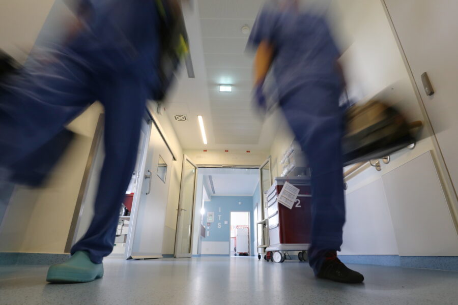 Zwei Personen in Krankenhauskleidung gehen fluraufwärts. Geräteschrank rechts.