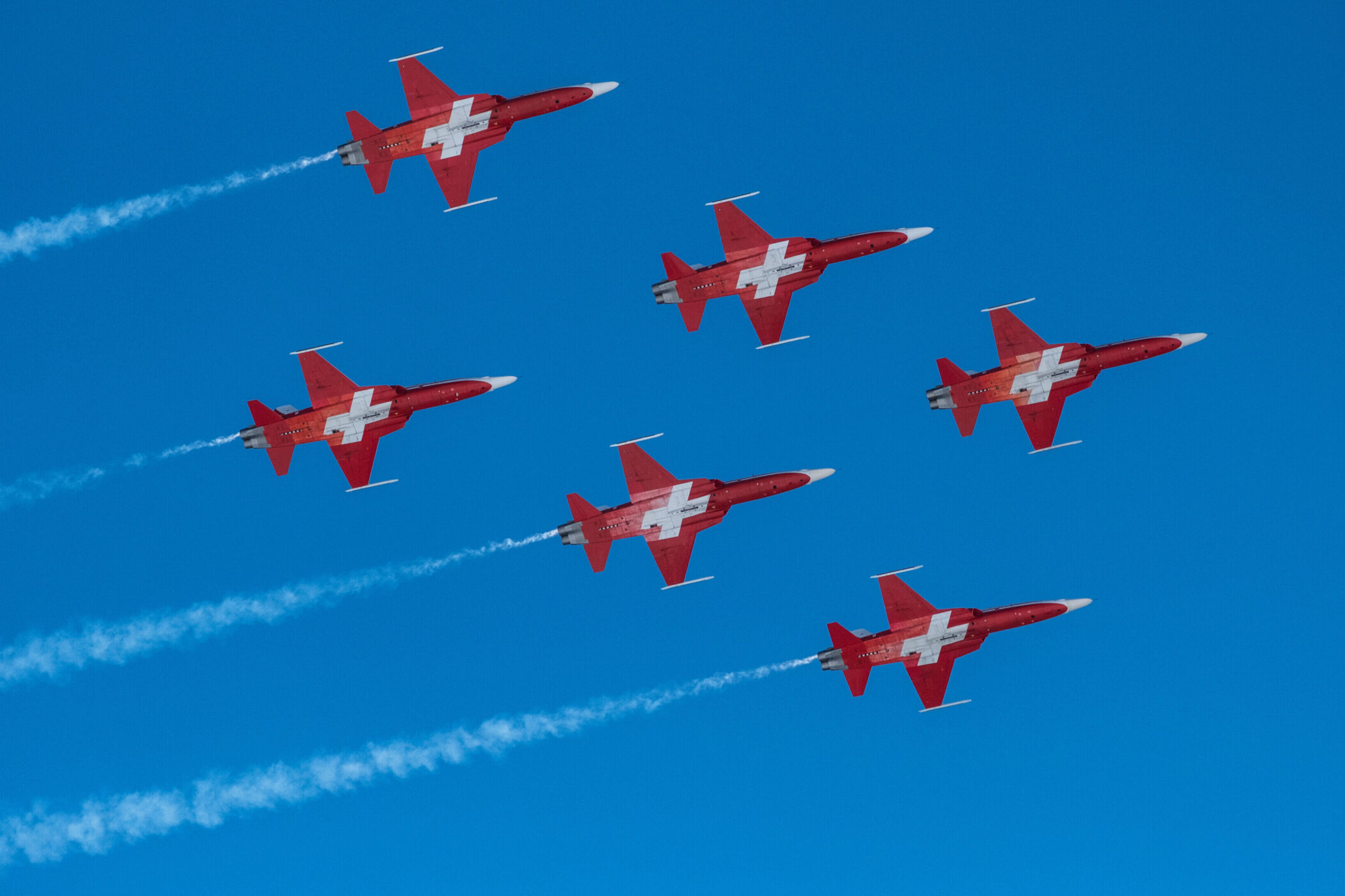 Sechs rote Flugzeuge mit weißen Kreuzen fliegen in Formation am blauen Himmel.