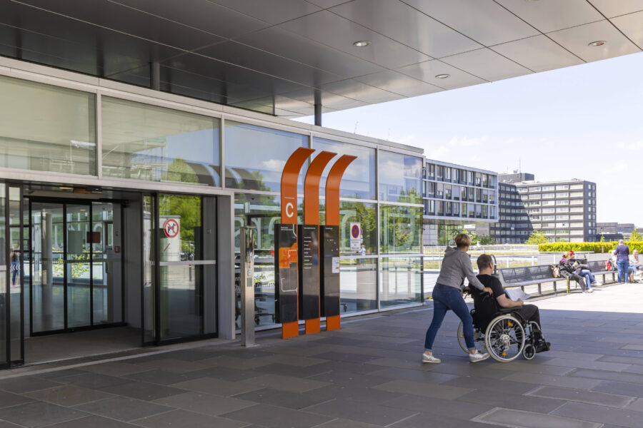 Glasgebäude mit Automat, Menschen im Hintergrund, Person im Rollstuhl vorne.