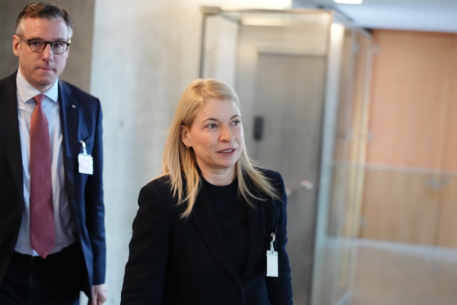 Zwei Personen gehen durch einen Flur, beide tragen Anzug und ID-Karte.