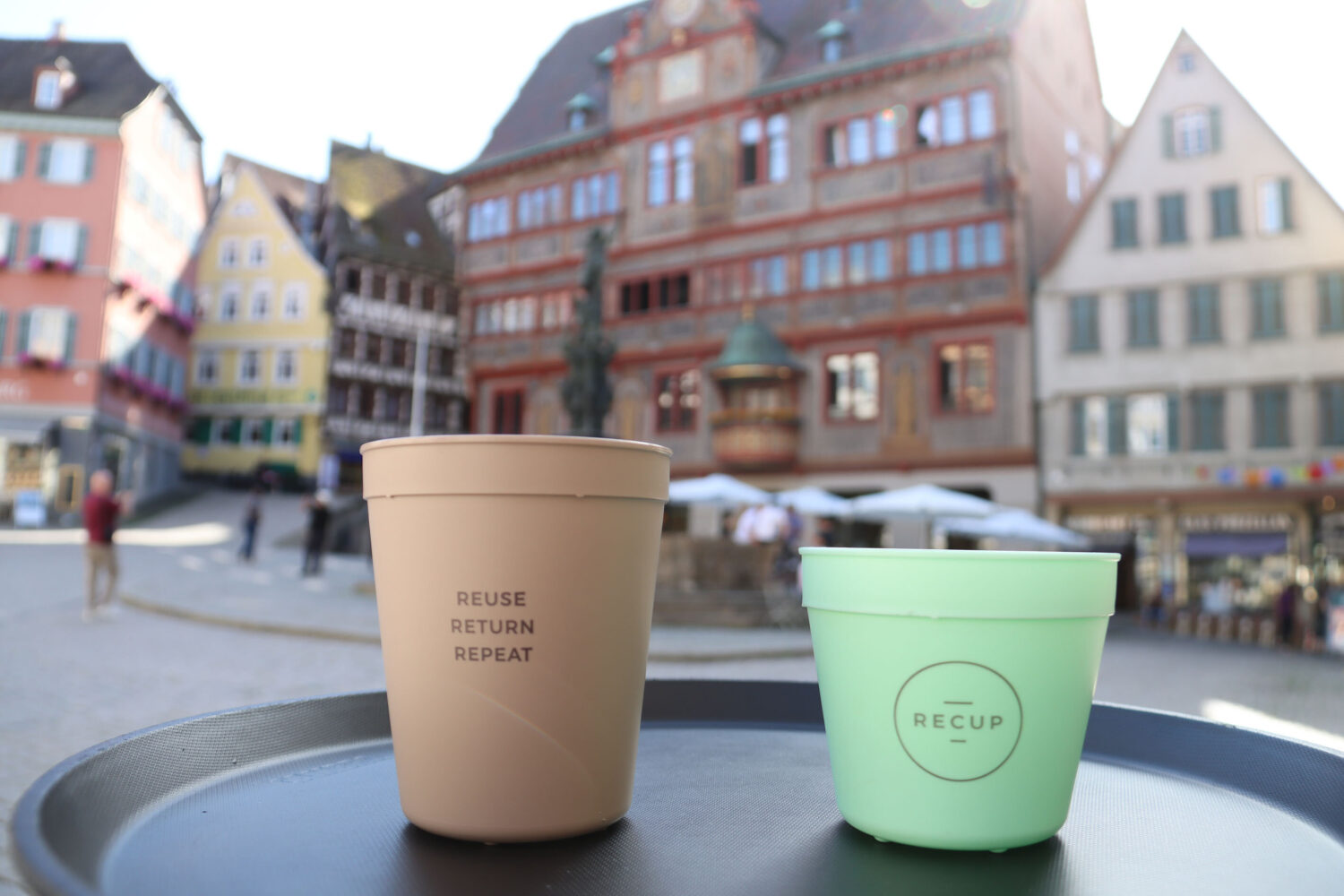 Zwei Becher auf Tablett, Stadtgebäude im Hintergrund. Becheraufschrift: „REUSE RETURN REPEAT“.