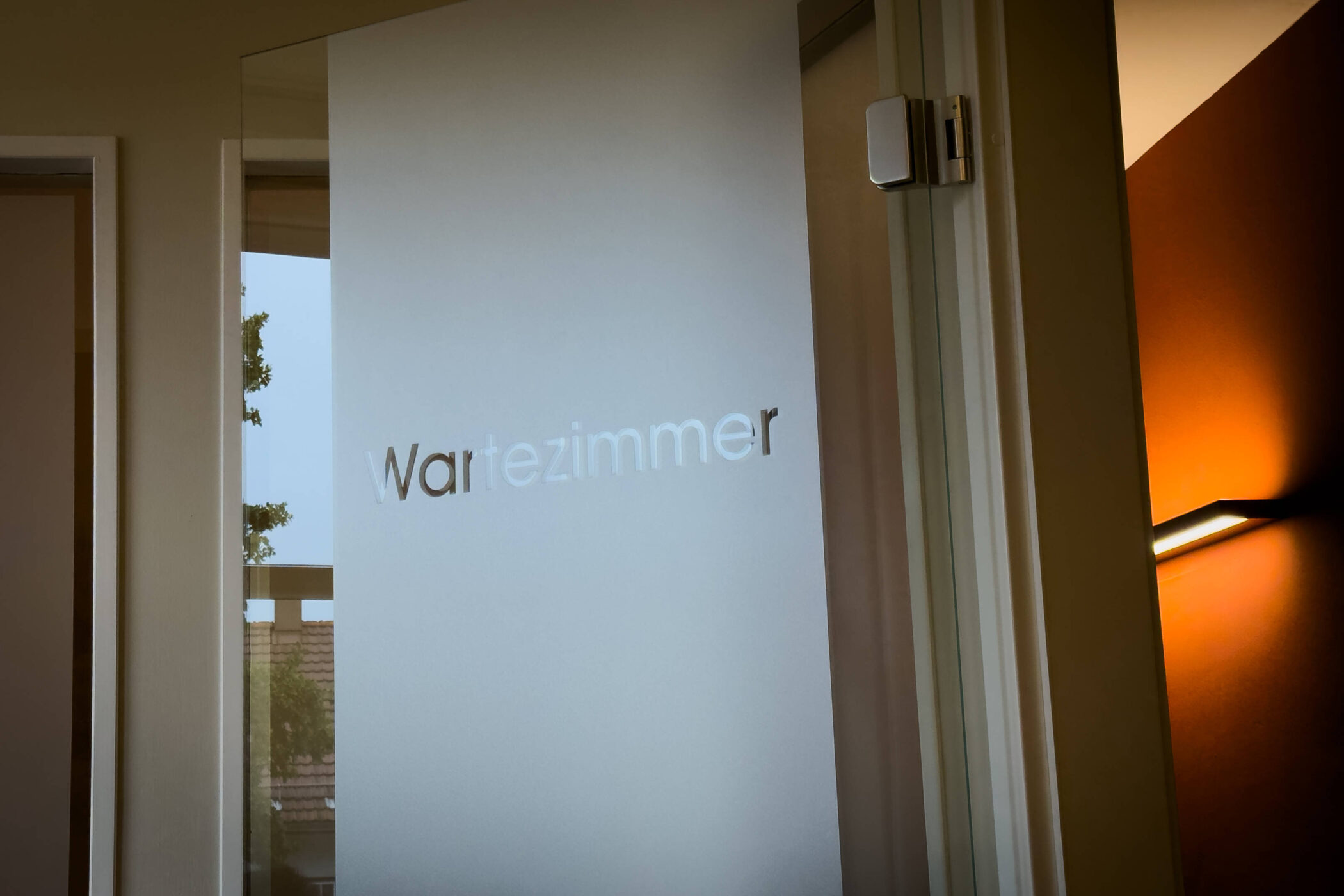 Glastür mit dem Text "Wartezimmer", dahinter orange beleuchteter Raum.
