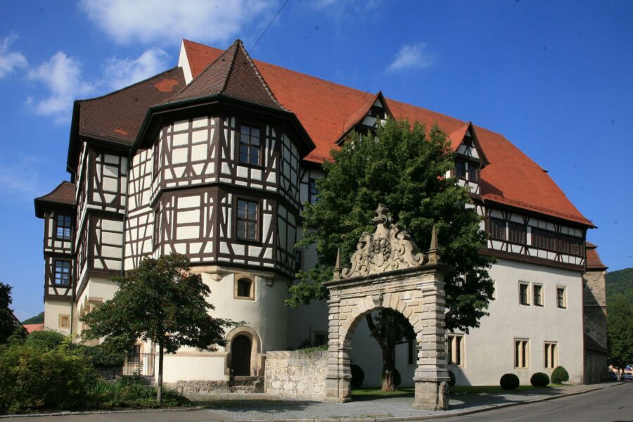 Fachwerkhaus mit rotem Dach und steinernem Torbogen davor unter blauem Himmel.