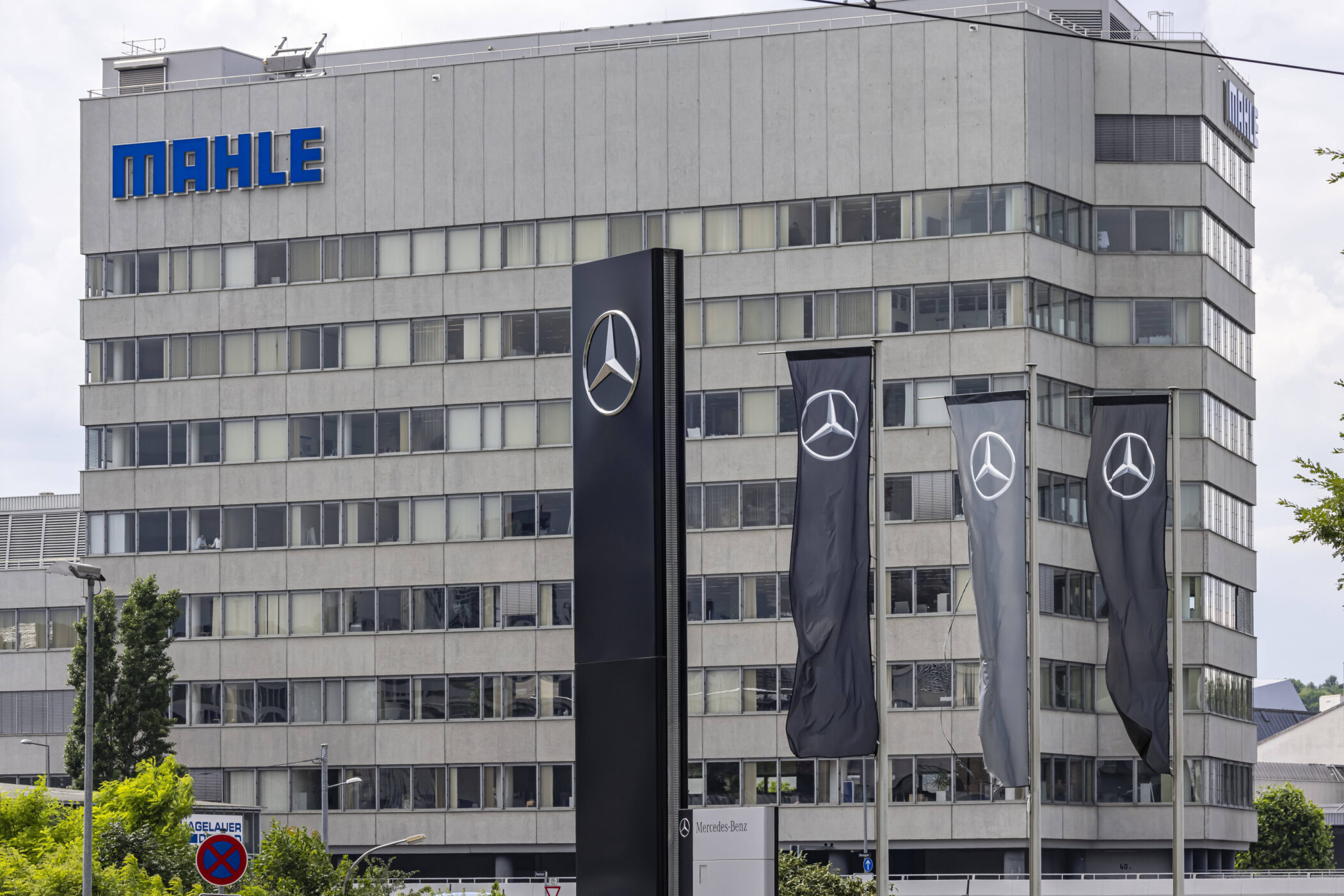 Graues Gebäude mit "MAHLE" Logo, Mercedes-Logo auf Flaggen und Schild.