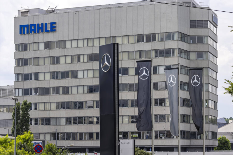 Graues Gebäude mit "MAHLE" Logo, Mercedes-Logo auf Flaggen und Schild.