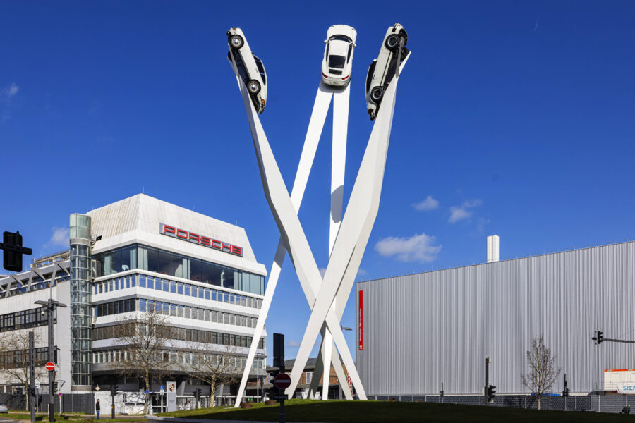 Drei weiße Autos auf hohen, geneigten Pfeilern; Gebäude mit Porsche-Logo daneben.
